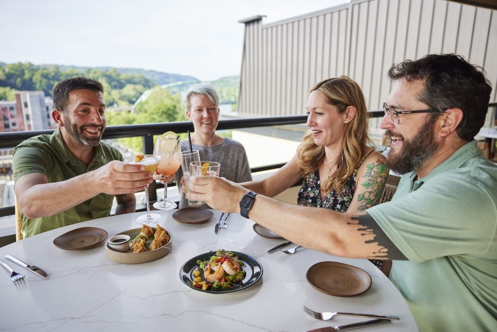 Four diners share appetizers and clink glasses dining outdoors at Thyme Rooftop Bar & Grille.